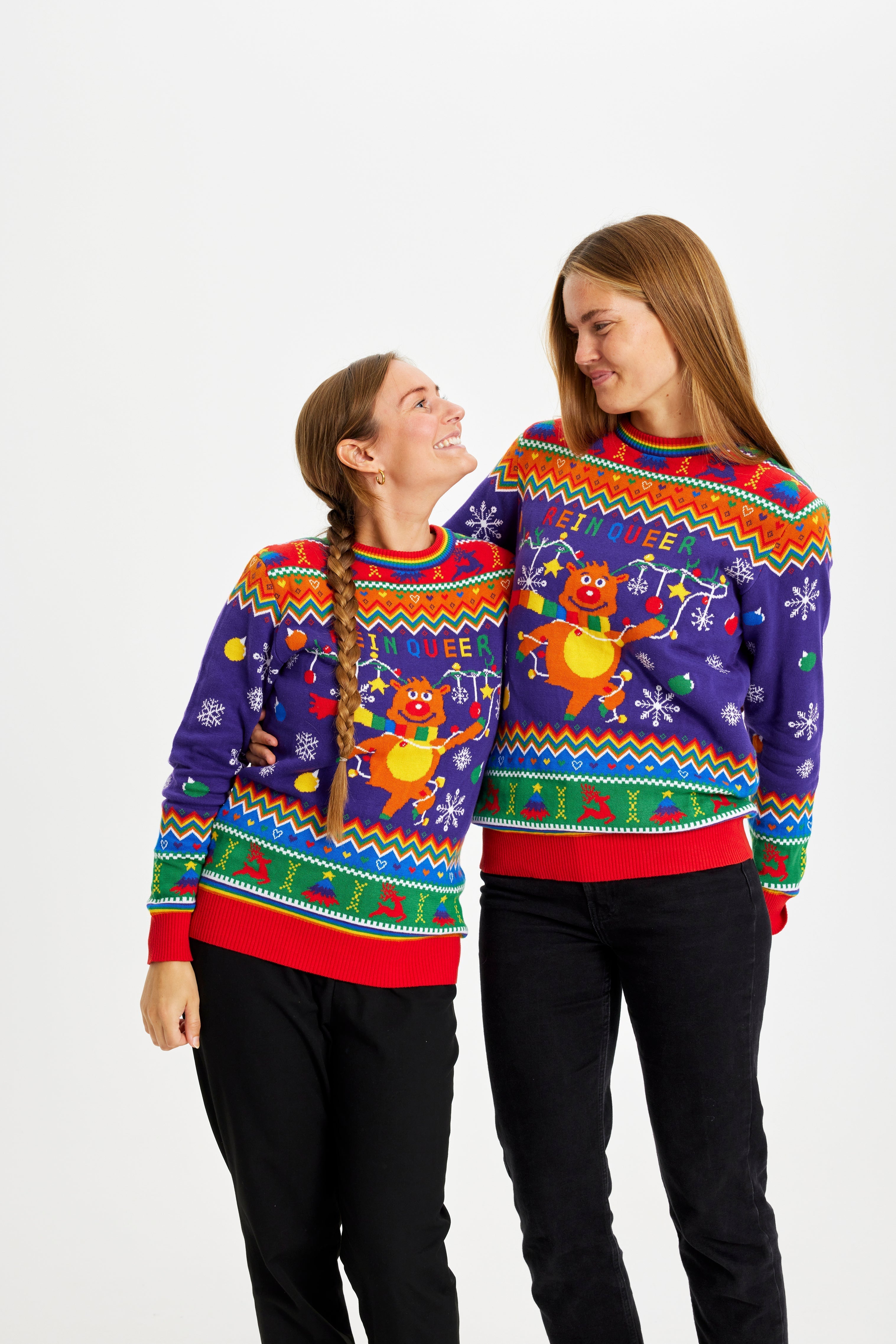 Twee vrouwen kijken elkaar aan terwijl ze een regenboogkleurige kersttrui dragen met de quote 